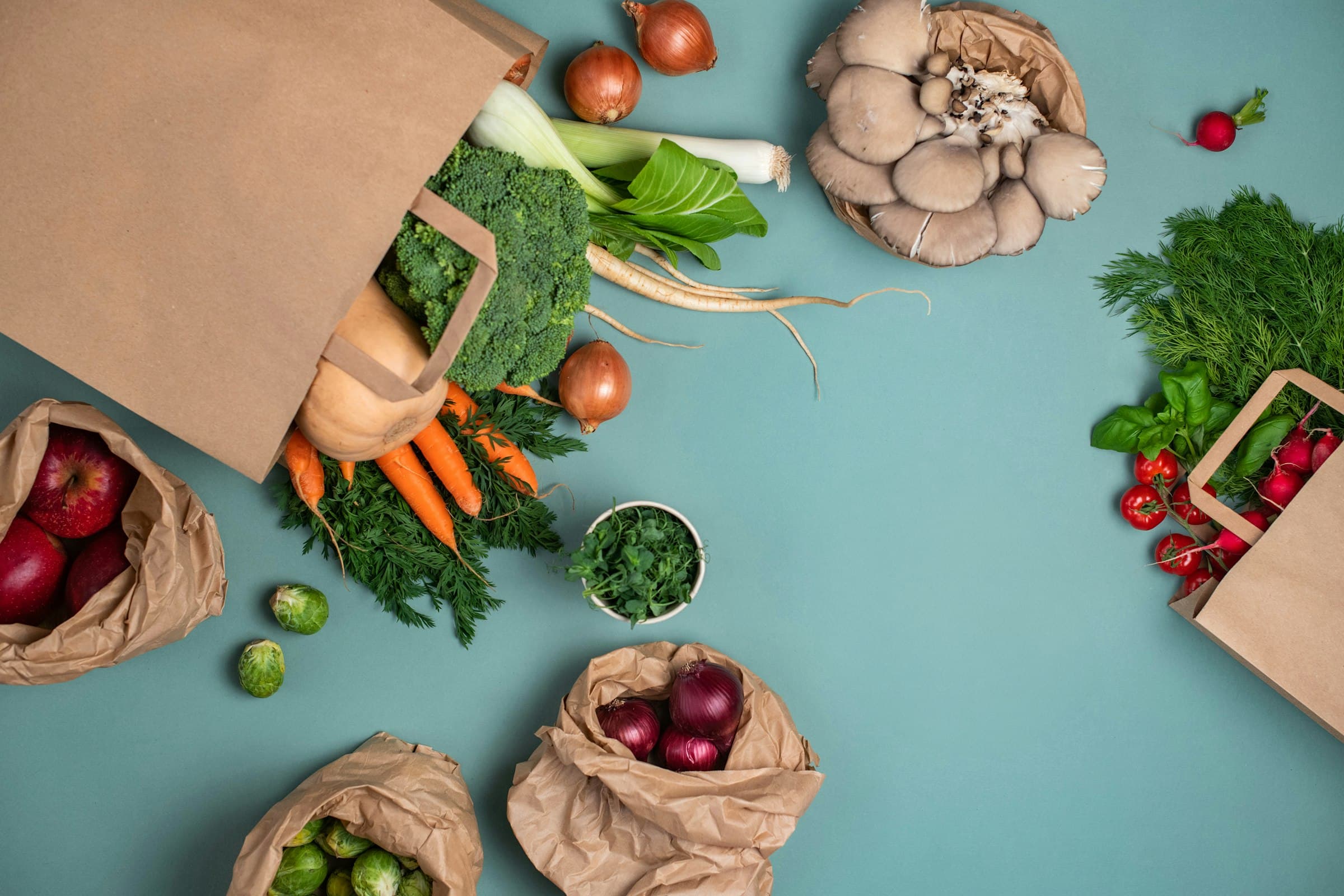 Brown paper grocery bags spilling broccoli, carrots, mushrooms, onions and herbs on a teal surface
