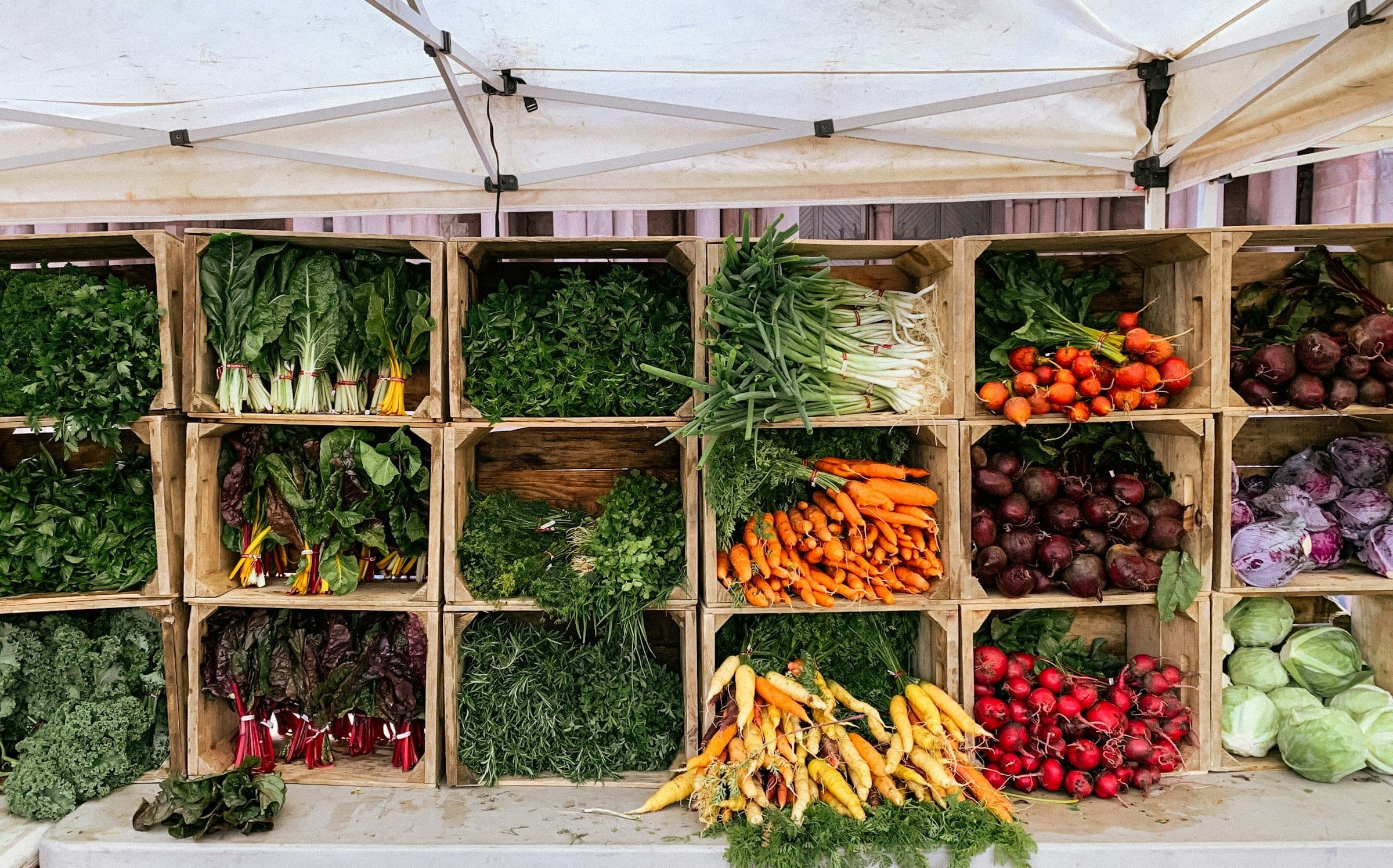 A market stall under a white canopy with wooden crates of greens, carrots, beets and cabbage
