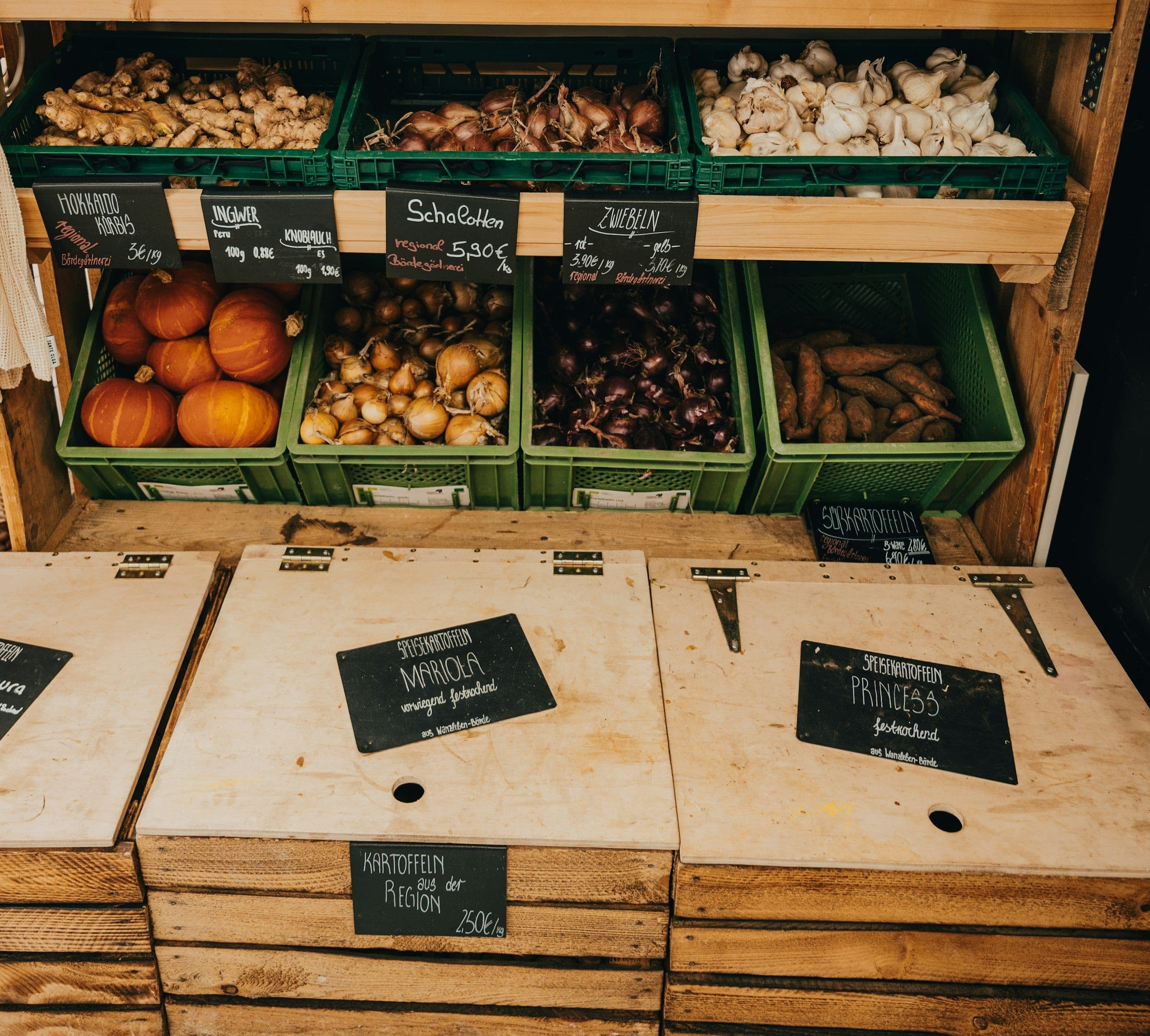 A shop interior with green crates of onions, pumpkins and garlic above wooden potato bins with chalkboard signs
