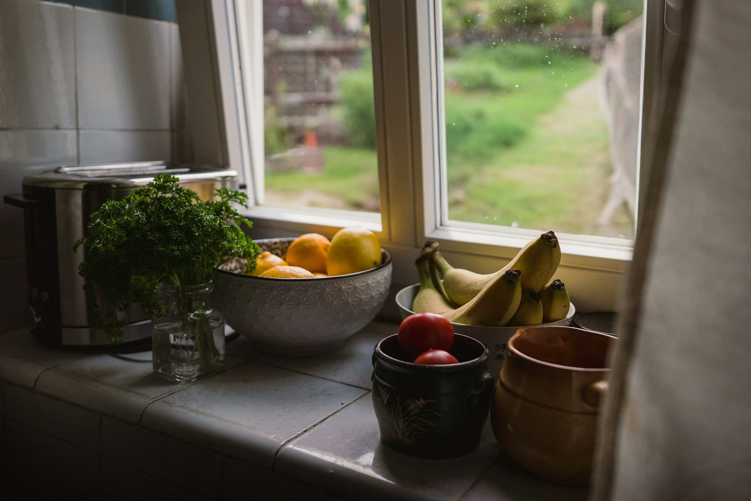 A kitchen windowsill with fresh herbs in a glass jar, a bowl of citrus and ripe bananas